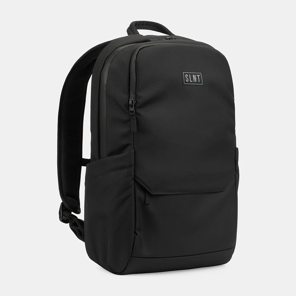 The SLNT Essentials Faraday Backpack by SLNT is black with padded shoulder straps, a top handle, front zip pocket, side pockets, and a small rectangular SLNT logo patch on the front, displayed against a plain light gray background.