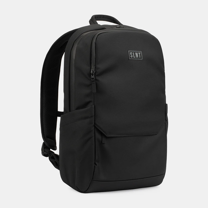 The SLNT Essentials Faraday Backpack by SLNT is black with padded shoulder straps, a top handle, front zip pocket, side pockets, and a small rectangular SLNT logo patch on the front, displayed against a plain light gray background.
