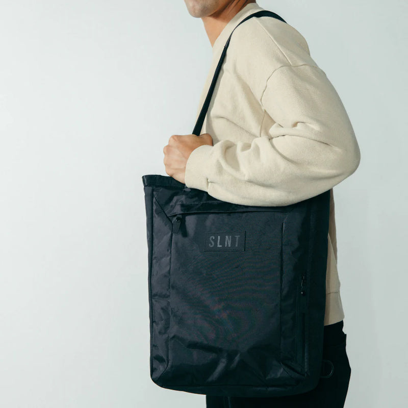 A person holds the SLNT E3 Faraday Tote Bag (17L) in black with the SLNT logo, slung over their shoulder, while wearing a light beige sweater against a plain light background.